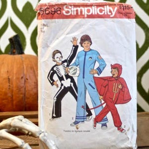 May include: A white Simplicity sewing pattern envelope, number 5696, featuring illustrations of three costume designs: a black and white skeleton, a light blue jumpsuit with a circular emblem, and a red devil outfit with a cape and a pitchfork.
