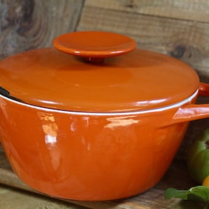 May include: Orange enamelled cast iron pot with a lid. The pot has two handles and a white rim. The lid has a knob on top.