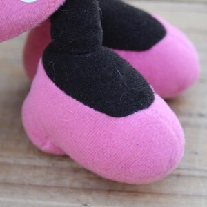 May include: Close-up of a pink plush toy's feet. The feet are shaped like shoes with black fabric on the top.