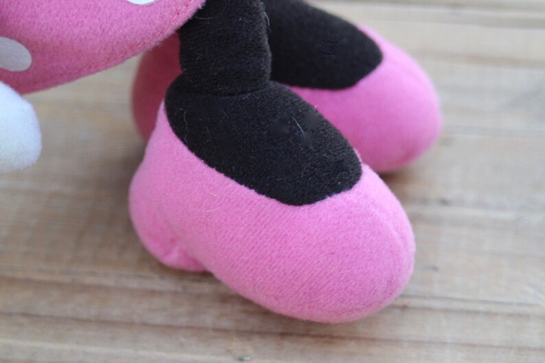 May include: Close-up of a pink plush toy's feet. The feet are shaped like shoes with black fabric on the top.