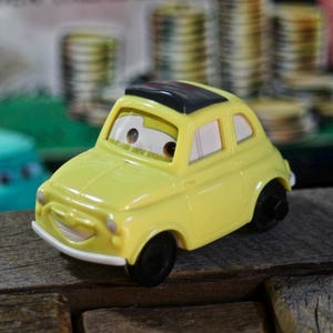 May include: A yellow toy car with a black roof and smiling face. The car has white-rimmed windows and black wheels. The toy car is sitting on a wooden surface.
