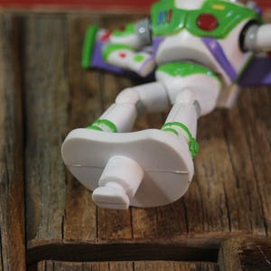 Puede incluir: Figura de acci&oacute;n de Buzz Lightyear blanca y verde, sobre una superficie de madera. El juguete tiene detalles verdes, alas moradas y un emblema rojo y verde en el pecho. Los pies de la figura son prominentes.