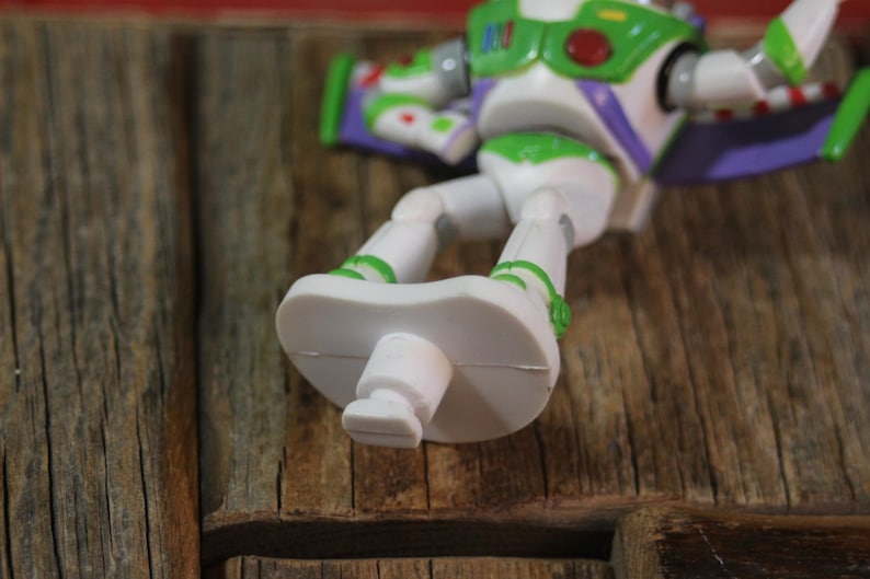 Puede incluir: Figura de acci&oacute;n de Buzz Lightyear blanca y verde, sobre una superficie de madera. El juguete tiene detalles verdes, alas moradas y un emblema rojo y verde en el pecho. Los pies de la figura son prominentes.