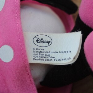 May include: Pink plush toy with white polka dots and a black face. The toy has a white tag with the text "Disney &copy;Disney Manufactured under license by Just Play, LLC 401 Fairway Drive, Deerfield Beach, FL 33441, USA"