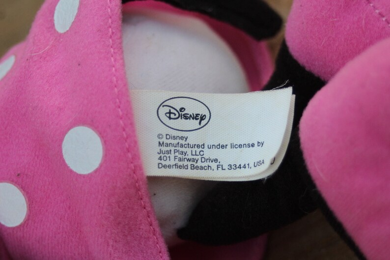 May include: Pink plush toy with white polka dots and a black face. The toy has a white tag with the text "Disney &copy;Disney Manufactured under license by Just Play, LLC 401 Fairway Drive, Deerfield Beach, FL 33441, USA"