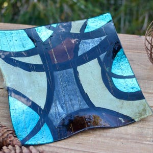 May include: A square glass serving tray with a geometric design in shades of blue, black, and brown. The tray has a wavy edge and a glossy finish.