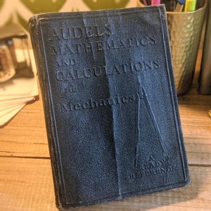 May include: A black hardcover book titled Audels Mathematics and Calculations for Mechanics. The cover features embossed text and a graphic of a drafting compass. The book is a ready reference guide with a textured surface and visible wear on the spine.