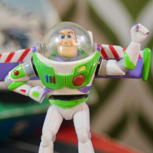 Puede incluir: Figura de acci&oacute;n de Buzz Lightyear, con casco transparente, traje espacial verde y blanco, y alas moradas. La figura est&aacute; posando con un brazo levantado, un dise&ntilde;o ic&oacute;nico de la franquicia Toy Story.