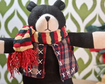 May include: A black plush bear wearing a red, green, and white striped scarf and a plaid vest. The bear has a white face and black eyes.