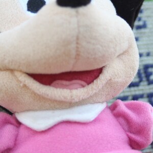 May include: A plush toy of a cartoon mouse wearing a pink dress with a white collar. The mouse has black ears and a black nose. The mouse is smiling and has a pink tongue sticking out.