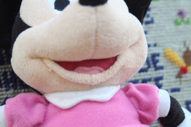 May include: A plush toy of a cartoon mouse wearing a pink dress with a white collar. The mouse has black ears and a black nose. The mouse is smiling and has a pink tongue sticking out.