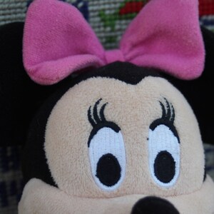 May include: A plush toy of a cartoon mouse with black ears and a pink bow. The mouse has large, white eyes with black pupils and eyelashes.