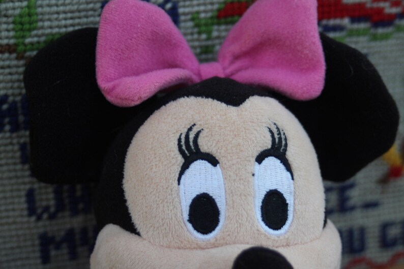 May include: A plush toy of a cartoon mouse with black ears and a pink bow. The mouse has large, white eyes with black pupils and eyelashes.