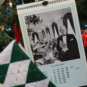 May include: A black and white photo of a man in a tuxedo standing in a dining room. The photo is on a calendar with the month of April 1964. The calendar is hanging on a Christmas tree with red and green ornaments.