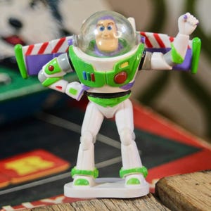 Puede incluir: Figura de acci&oacute;n de Buzz Lightyear, con casco transparente, traje espacial verde y blanco, y alas moradas y rojas. La figura posa con un brazo levantado, de pie sobre una base blanca. El fondo incluye un juego de mesa y una superficie de madera.
