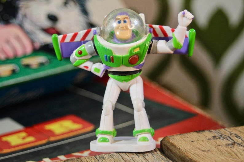 Puede incluir: Figura de acci&oacute;n de Buzz Lightyear, con casco transparente, traje espacial verde y blanco, y alas moradas y rojas. La figura posa con un brazo levantado, de pie sobre una base blanca. El fondo incluye un juego de mesa y una superficie de madera.