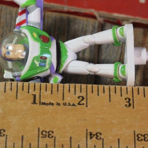 Puede incluir: Figura de acci&oacute;n de Buzz Lightyear, con un traje espacial verde y blanco, detalles morados y un casco transparente. La figura mide aproximadamente 7,6 cm de largo, junto a una regla de madera.