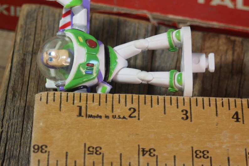 Puede incluir: Figura de acci&oacute;n de Buzz Lightyear, con un traje espacial verde y blanco, detalles morados y un casco transparente. La figura mide aproximadamente 7,6 cm de largo, junto a una regla de madera.