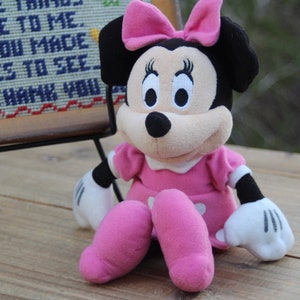 May include: A pink and black plush toy of Minnie Mouse wearing a pink dress and a pink bow. The toy is sitting on a wooden surface.
