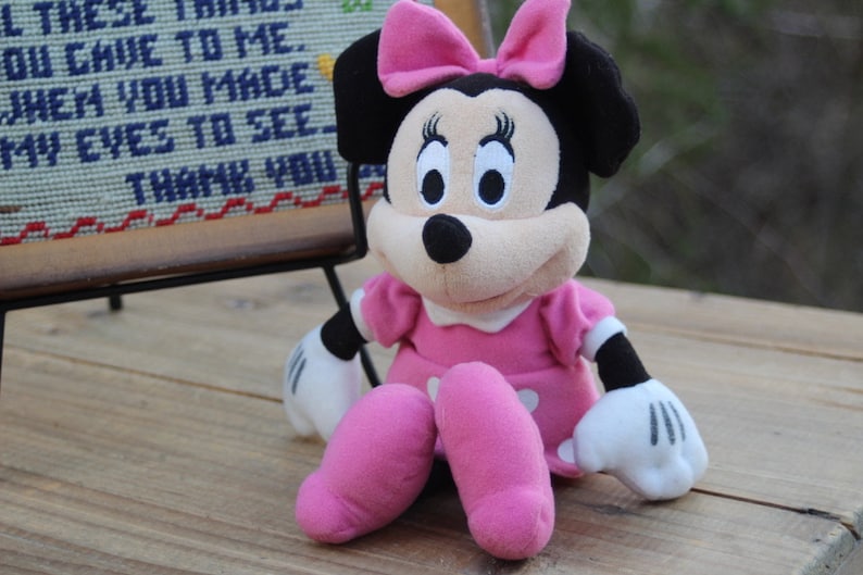 May include: A pink and black plush toy of Minnie Mouse wearing a pink dress and a pink bow. The toy is sitting on a wooden surface.