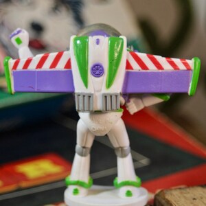 Puede incluir: Una figura de pl&aacute;stico de Buzz Lightyear, vista desde atr&aacute;s, con un cuerpo blanco, alas verdes y moradas, y detalles a rayas rojas y blancas. La figura est&aacute; sobre una base blanca. Se ven las palabras "MADE IN CHINA".