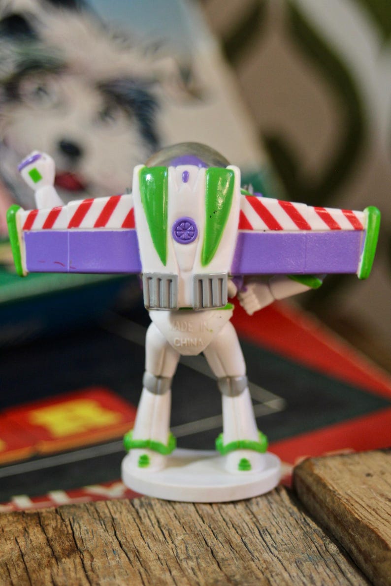 Puede incluir: Una figura de pl&aacute;stico de Buzz Lightyear, vista desde atr&aacute;s, con un cuerpo blanco, alas verdes y moradas, y detalles a rayas rojas y blancas. La figura est&aacute; sobre una base blanca. Se ven las palabras "MADE IN CHINA".