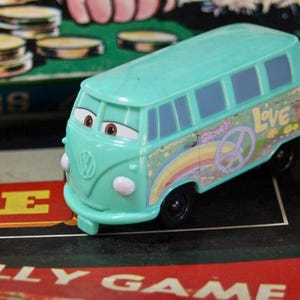 May include: A teal toy bus with cartoon eyes and a peace sign design. The bus has a rainbow and the word "Love" on the side. The toy is on a game board.
