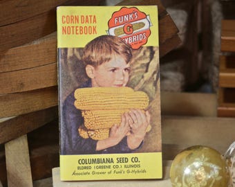 May include: A yellow and red notebook with the text "CORN DATA NOTEBOOK" and "FUNK'S G HYBRIDS" on the cover. A young boy is holding a large ear of corn in front of him. The notebook is from Columbiana Seed Co. in Eldred, Illinois.