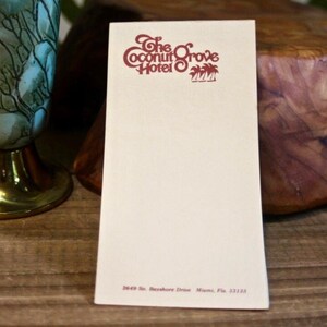 May include: A rectangular off-white notepad featuring the dark red text The Coconut Grove Hotel with a palm tree graphic at the top and the address 2649 So. Bayshore Drive, Miami, Fla. 33133 printed in small dark red font at the bottom.