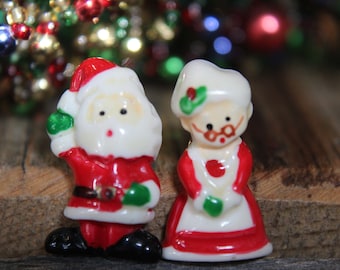 May include: A pair of ceramic Christmas ornaments depicting Santa Claus and Mrs. Claus. Santa is wearing a red suit with a white trim and a white beard. Mrs. Claus is wearing a red dress with a white apron and a white bonnet.