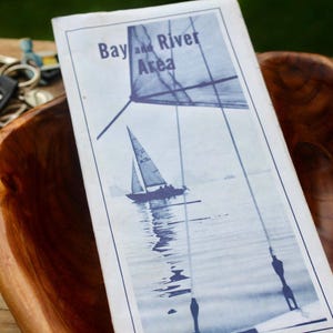 May include: A white rectangular brochure with blue ink text reading Bay and River Area, featuring a graphic of a sailboat on water. The brochure is placed inside a polished, natural-toned wooden bowl with a visible grain pattern and smooth curves.