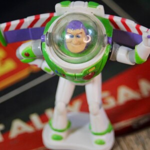 Puede incluir: Una figura de acci&oacute;n de Buzz Lightyear, con un traje espacial blanco y verde con un casco transparente. La figura tiene detalles en morado y alas a rayas rojas y blancas. El juguete est&aacute; sobre una base blanca.