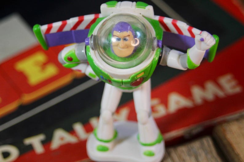 Puede incluir: Una figura de acci&oacute;n de Buzz Lightyear, con un traje espacial blanco y verde con un casco transparente. La figura tiene detalles en morado y alas a rayas rojas y blancas. El juguete est&aacute; sobre una base blanca.