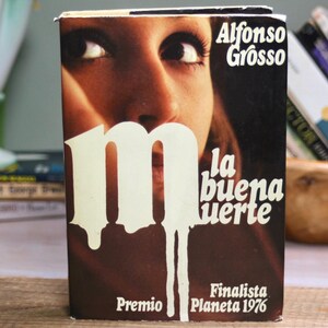 May include: A hardcover book titled La buena muerte by Alfonso Grosso, featuring a dark brown cover with a large white letter M and a close-up photograph of a person's eyes. The text Premio Planeta 1976 Finalista is printed at the bottom.