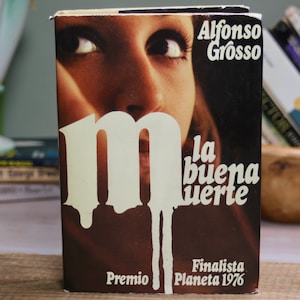 May include: A brown paperback book with a white "M" on the cover. The title is "La buena muerte" by Alfonso Grosso. The book is a finalist for the Planeta 1976 award.