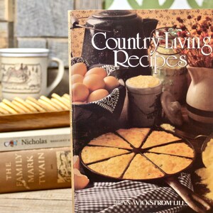 May include: A still life featuring a cookbook titled "Country Living Recipes" with a vintage aesthetic. The cover displays a rustic scene with food items like eggs, a pie, and a coffee pot. A stack of books and a mug are in the background.