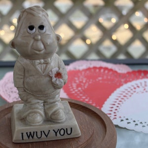 May include: A ceramic figurine of a man with large eyes and a flower in his hand. The figurine is standing on a base that says "I WUV YOU".