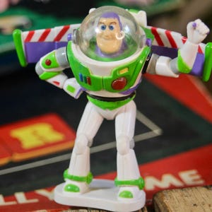 Puede incluir: Figura de acci&oacute;n de Buzz Lightyear, con un traje espacial blanco y verde y un casco transparente. La figura tiene un bot&oacute;n rojo en el pecho y alas con rayas rojas y blancas. El juguete est&aacute; sobre una base blanca.