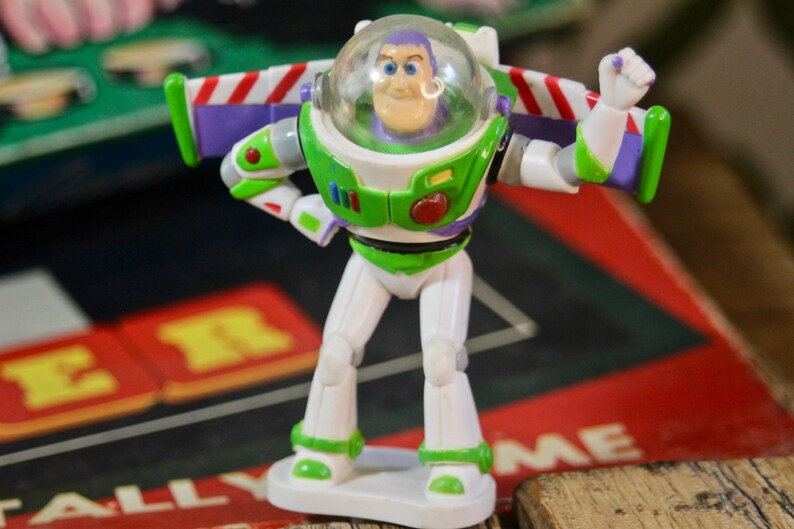 Puede incluir: Figura de acci&oacute;n de Buzz Lightyear, con un traje espacial blanco y verde y un casco transparente. La figura tiene un bot&oacute;n rojo en el pecho y alas con rayas rojas y blancas. El juguete est&aacute; sobre una base blanca.