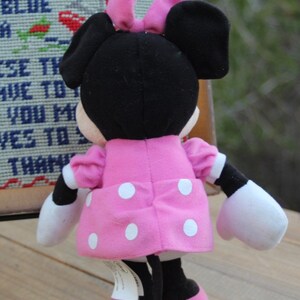 May include: A pink and white polka dot dress with a black bow on top of the head. The plush toy is a black and white Minnie Mouse.