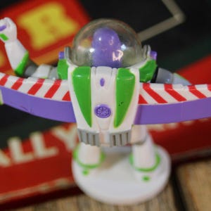 Puede incluir: Figura de acci&oacute;n de Buzz Lightyear, con un traje espacial blanco y verde con detalles morados y rojos. La figura tiene un casco transparente y est&aacute; posando con los brazos extendidos, las alas extendidas. El juguete est&aacute; sobre una base blanca.