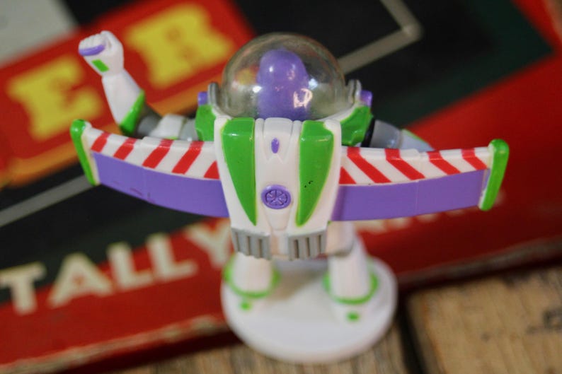 Puede incluir: Figura de acci&oacute;n de Buzz Lightyear, con un traje espacial blanco y verde con detalles morados y rojos. La figura tiene un casco transparente y est&aacute; posando con los brazos extendidos, las alas extendidas. El juguete est&aacute; sobre una base blanca.