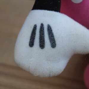 May include: Close-up of a white plush glove with three black stitched fingers. The glove is part of a plush toy.