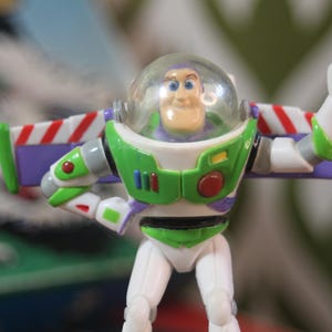 Puede incluir: Figura de acci&oacute;n de Buzz Lightyear, con un traje espacial blanco y verde con detalles morados y rojos. La figura tiene un casco transparente y alas extendidas. El juguete est&aacute; posando con un pu&ntilde;o cerrado.