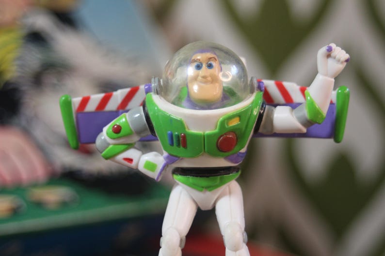 Puede incluir: Figura de acci&oacute;n de Buzz Lightyear, con un traje espacial blanco y verde con detalles morados y rojos. La figura tiene un casco transparente y alas extendidas. El juguete est&aacute; posando con un pu&ntilde;o cerrado.