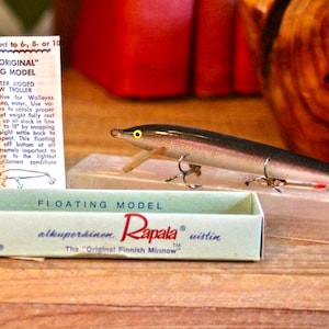 May include: A Rapala Original Floating Model fishing lure with a black back and silver sides, featuring a yellow eye and two treble hooks. It is displayed with its light blue cardboard box and a white instruction paper with black text.