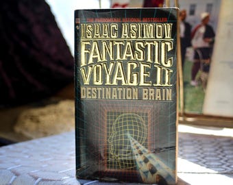 May include: A paperback book titled Fantastic Voyage II: Destination Brain by Isaac Asimov. The black cover features gold embossed lettering and a yellow wireframe graphic of a human head profile with a light beam pointing toward the brain.