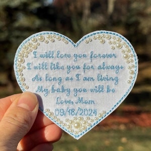May include: A white heart-shaped fabric patch with blue embroidery. The patch has a message that reads "I will love you forever I will like you for always As long as I am living My baby you will be Love, Mom 09/18/2024". The patch is decorated with white pearl-like beads around the edges.