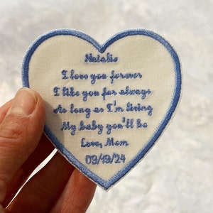 May include: A white heart-shaped patch with blue embroidery. The patch says "Natalie I love you forever I like you for always As long as I'm living My baby you'll be Love, Mom 09/19/24".
