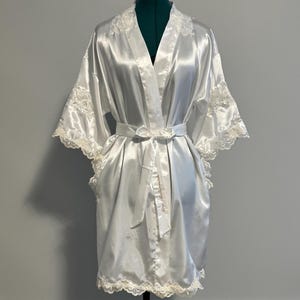 May include: A white satin robe with lace trim. The robe has a long sleeve and a sash tie. The robe is displayed on a mannequin.